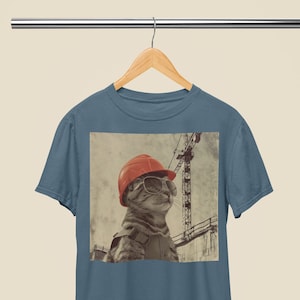 Puede incluir: Una camiseta azul con una imagen de estilo vintage de un gato con un casco y gafas de sol delante de una grúa de construcción.