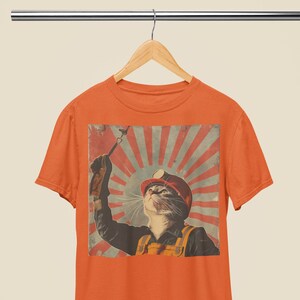 Puede incluir: Camiseta naranja con un gráfico de estilo vintage de un gato con un casco de minero y sosteniendo un pico. El gráfico está sobre un fondo de sol rojo y blanco.