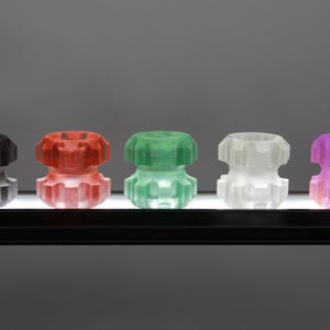 Könnte beinhalten: Fünf kleine, durchsichtige Dekorationsobjekte in verschiedenen Farben. Die Objekte sind übereinander gestapelt und haben ein geometrisches Design. Die Farben sind Schwarz, Rot, Grün, Weiß und Lila.