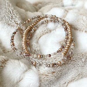 May include: A set of three beaded bracelets in shades of brown, beige, and clear. The bracelets are layered on top of each other, creating a multi-layered look.