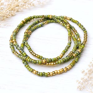 May include: A set of three layered bracelets made from small round beads in shades of olive green, mottled sage, and metallic gold. The beads are arranged in a repeating pattern, alternating between solid green, speckled green, and shiny gold accents.