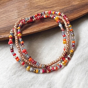 May include: Three beaded bracelets in shades of red, orange, yellow, and silver. The bracelets are made with small, colorful beads and a gold-colored metal. The bracelets are arranged on a wooden surface.