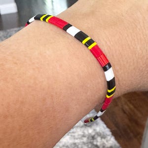 Pode incluir: Um bracelete de contas vermelhas, pretas, brancas e amarelas. O bracelete é feito de pequenas contas quadradas.