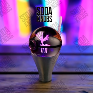 Peut inclure: Un bouton de soda personnalisé avec un design sphérique transparent. Le bouton présente un motif rose en forme d'arbre et des lignes verticales, avec le texte "SODA KNOBS" au-dessus. Le bouton repose sur une base argentée.