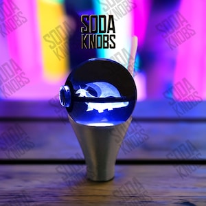 Könnte beinhalten: Ein blau-silberner Soda-Knauf mit einem detaillierten Design im Inneren einer transparenten Kugel. Die Kugel zeigt eine weiße Illustration einer Kreatur. Der Text "SODA KNOBS" wird über dem Knauf angezeigt.