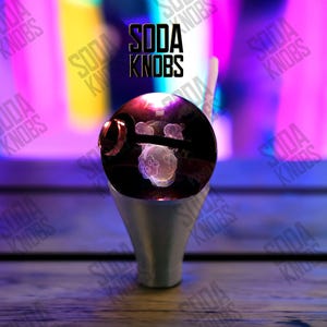 Peut inclure: Un bouton de soda unique avec un dessus sphérique transparent présentant un motif de méduse. Le bouton a une base argentée et le texte "SODA KNOBS" est affiché en noir. L'arrière-plan est un mélange flou de violet, de jaune et de rose.