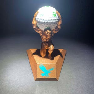 May include: A bronze statue of a man holding a golf ball aloft. The base of the statue is a triangular piece of wood with a blue eagle design.