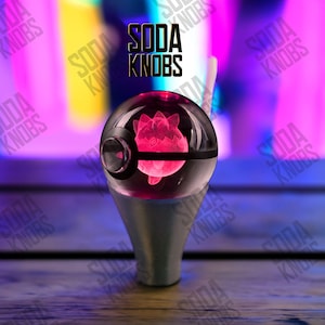 Peut inclure: Un bouton de soda sphérique transparent avec un personnage rose et lumineux à l'intérieur. Le bouton a une bande noire et une base argentée. Le texte "SODA KNOBS" est affiché en noir au-dessus du bouton.