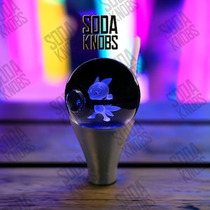 Peut inclure: Un pommeau de levier de vitesse personnalisé avec un personnage Pokémon bleu et lumineux à l'intérieur d'une sphère transparente. Le pommeau repose sur une base argentée. Le texte "SODA KNOBS" est affiché.