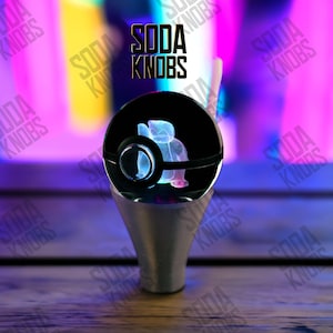 Könnte beinhalten: Ein schwarzer und silberner Soda-Knopf mit dem Text "SODA KNOBS". Der Knopf hat ein klares, rundes Design mit einer blau-rosa Grafik. Der Sockel ist silberfarben und kegelförmig.