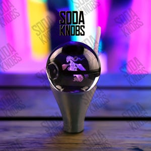 Könnte beinhalten: Ein klarer, kugelförmiger Schalthebelknauf mit schwarzem Sockel und silbernem Schaft. Der Knauf hat ein lila, 3D-geätztes Design einer mythischen Kreatur. Der Text "SODA KNOBS" wird in Schwarz angezeigt.