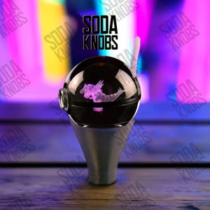 Könnte beinhalten: Ein schwarzer, kugelförmiger Soda-Knopf mit silbernem Sockel. Der Knopf zeigt ein rosa, geätztes Drachendesign und den Text "SODA KNOBS" in Schwarz. Der Hintergrund ist eine verschwommene Mischung aus Lila, Rosa und Gelb.