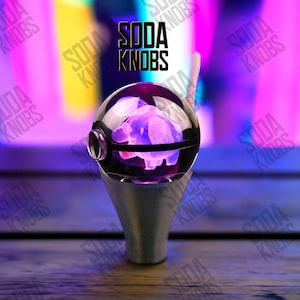 Peut inclure: Un bouton de soda décoratif avec un dessus sphérique transparent représentant un cochon violet. Le bouton a une base argentée et le texte "SODA KNOBS" est visible en arrière-plan. Le thème général est la fantaisie.