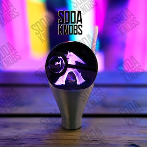 May include: A custom soda knob with a black, spherical top featuring a white, glowing design. The knob is mounted on a silver-coloured base. The text "SODA KNOBS" is displayed in black above the knob.