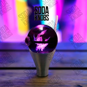 Peut inclure: Un bouton de verre décoratif avec un motif violet et lumineux à l'intérieur, monté sur une base argentée. Le texte "SODA KNOBS" est affiché au-dessus du bouton en noir. L'arrière-plan est un mélange flou de violet, de rose et de jaune.
