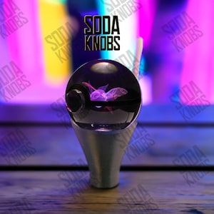 Puede incluir: Un pomo de soda decorativo con una parte superior esférica transparente que presenta un diseño púrpura brillante. El pomo está montado sobre una base plateada. El texto "SODA KNOBS" se muestra encima del pomo.