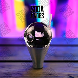 Könnte beinhalten: Ein schwarzer und klarer Soda-Knauf mit einem Cartoon-Charakter-Design. Der Knauf hat einen silbernen Sockel und der Text "SODA KNOBS" wird in Schwarz über dem Knauf angezeigt.