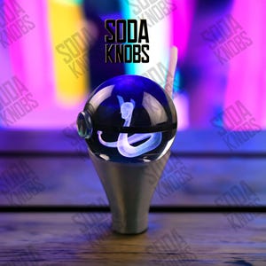 Op de afbeelding: Een heldere, bolvormige sodaknop met een gloeiend wit slangontwerp aan de binnenkant. De knop staat op een zilverkleurige basis. De tekst "SODA KNOBS" staat in het zwart boven de knop.