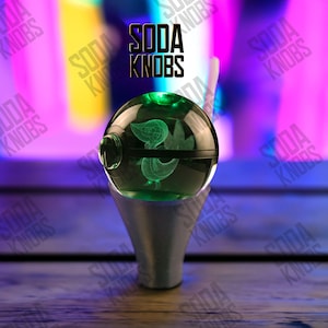 Peut inclure: Un bouton de soda sphérique vert avec un motif gravé, reposant sur une base argentée. Le bouton est éclairé d'une lumière verte. Le texte "SODA KNOBS" est affiché au-dessus du bouton.