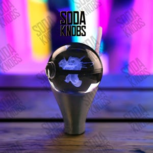 Peut inclure: Un bouton de soda sphérique transparent avec un motif de méduse bleu à l'intérieur, monté sur une base argentée. Le texte "SODA KNOBS" est affiché au-dessus du bouton en lettres noires.