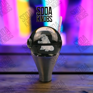 Könnte beinhalten: Ein schwarzer und klarer Soda-Knopf aus Glas mit einem weißen, geätzten Design eines Zeichentrickcharakters. Der Knopf ist auf einem silberfarbenen Sockel montiert. Der Text "SODA KNOBS" wird in Schwarz angezeigt.