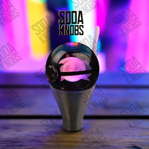 Könnte beinhalten: Ein individueller Soda-Knopf mit einem transparenten, kugelförmigen Oberteil mit einem rosa, leuchtenden Quallen-Design. Der Knopf ist auf einem silberfarbenen Sockel montiert. Der Text "SODA KNOBS" wird in Schwarz über dem Knopf angezeigt.