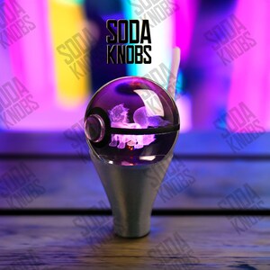 Op de afbeelding: Een heldere, bolvormige sodaknop met een paars interieur, met een gloeiend wezenontwerp. De knop staat op een zilveren voet. De tekst "SODA KNOBS" wordt boven de knop weergegeven.