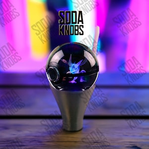 Peut inclure: Un bouton de soda décoratif avec un dessus sphérique transparent présentant un motif de personnage bleu et violet. Le bouton est monté sur une base argentée en forme de cône. Le texte "SODA KNOBS" est visible en arrière-plan.