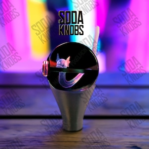 Peut inclure: Un bouton de soda noir et transparent avec un dessin de personnage de dessin animé. Le bouton a une base argentée et un design rose et blanc. Le texte "SODA KNOBS" est visible.
