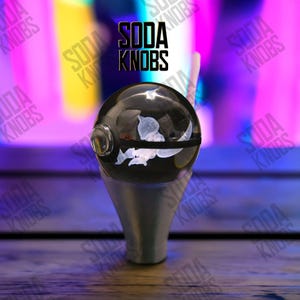 Puede incluir: Un pomo de soda negro y transparente con un diseño floral blanco y una base plateada. El texto "SODA KNOBS" se muestra en negro encima del pomo. El fondo es una mezcla borrosa de morado, amarillo y rosa.
