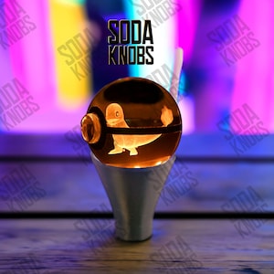 Puede incluir: Un pomo de soda esférico y brillante con un diseño de Charmander. El pomo está sobre una base plateada, con el texto "SODA KNOBS" arriba. El fondo es una mezcla borrosa de morado, amarillo y azul.