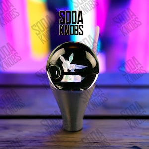 Peut inclure: Un bouton de soda en verre noir avec un motif de lapin blanc à l'intérieur. Le bouton repose sur une base argentée. Le texte "SODA KNOBS" est affiché au-dessus du bouton en noir, dans une police stylisée.