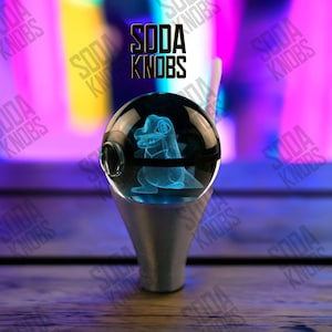 Könnte beinhalten: Ein klarer Soda-Knauf aus Glas mit einem blau leuchtenden Dinosaurier-Design im Inneren. Der Knauf ist auf einem silberfarbenen Sockel montiert. Der Text "SODA KNOBS" ist darüber zu sehen.