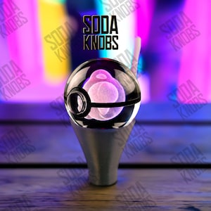 Könnte beinhalten: Ein durchsichtiger Glasflaschenverschluss mit einem schwarz-weißen Design, der ein rosa, kugelförmiges Design im Inneren aufweist. Der Verschluss ist auf einem silberfarbenen Sockel montiert. Der Text "SODA KNOBS" ist im Hintergrund sichtbar.
