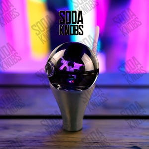 May include: A unique soda knob with a black, spherical top and a silver base. The knob features a purple design inside the sphere and the text "SODA KNOBS" in black. The background is a blurred mix of purple, yellow, and pink.