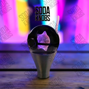 Op de afbeelding: Een zwarte en zilveren sodaknop met een gloeiend, paars, geometrisch wezenontwerp in een heldere bol. De tekst "SODA KNOBS" staat boven de knop. De achtergrond is een vage mix van paars, geel en roze.