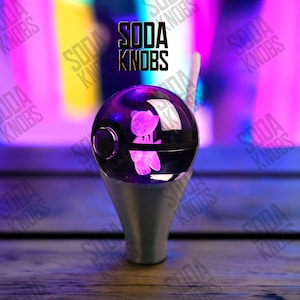 Op de afbeelding: Een heldere, bolvormige sodaknop met een gloeiende roze teddybeer erin. De knop staat op een zilveren voet. De tekst "SODA KNOBS" staat in het zwart boven de knop tegen een kleurrijke achtergrond.