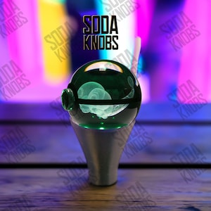 Könnte beinhalten: Ein grüner Glas-Sodaknopf mit silbernem Sockel, der ein leuchtendes Design im Inneren aufweist. Der Text "SODA KNOBS" wird in Schwarz über dem Knopf angezeigt. Der Knopf wird von grünen Lichtern beleuchtet.