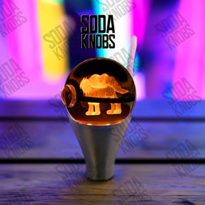 Peut inclure: Un bouton de soda sphérique transparent avec un motif de personnage orange éclairé à l'intérieur. Le bouton est posé sur une base argentée. Le texte "SODA KNOBS" est affiché en noir au-dessus du bouton sur un fond coloré.