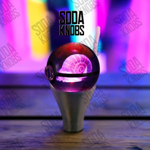 Peut inclure: Un bouton de soda sphérique et lumineux, avec un schéma de couleurs violet et rouge, présentant un motif en spirale à l'intérieur. Le bouton est monté sur une base argentée. Le texte "SODA KNOBS" est affiché au-dessus.