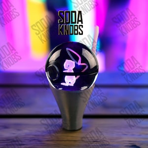 Peut inclure: Un bouton de soda sphérique transparent avec un motif de personnage de dessin animé gravé en 3D, monté sur une base argentée. Le texte "SODA KNOBS" est affiché au-dessus du bouton. L'arrière-plan est un mélange flou de violet, de rose et de jaune.