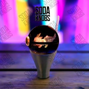 Puede incluir: Un pomo de soda decorativo con un orbe negro que presenta un diseño de conejo brillante. El pomo tiene una base plateada y detalles dorados. El texto "SODA KNOBS" se muestra encima del pomo.