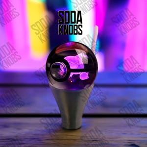 Puede incluir: Un pomo de soda esférico transparente con un interior púrpura, que presenta un diseño de personaje brillante. El pomo está montado sobre una base plateada. El texto "SODA KNOBS" se muestra encima del pomo.