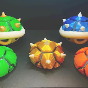 May include: A collection of colorful, 3D-printed Koopa Troopa shells in green, blue, orange, and gold. Each shell features a segmented design with white and yellow accents, and a ring on the bottom. The shells are inspired by the Super Mario video game series.