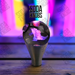 Peut inclure: Un bouton de soda décoratif avec un dessus sphérique transparent présentant un motif d'hippocampe rose et violet. Le bouton a une base argentée et le texte "SODA KNOBS" est affiché au-dessus.