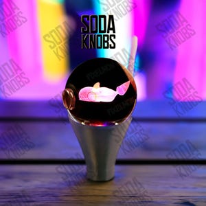 Könnte beinhalten: Ein dekorativer Soda-Knopf mit einem Wal-Design in einer klaren, runden Glaskugel. Der Knopf hat einen silbernen Sockel. Der Text "SODA KNOBS" ist sichtbar.