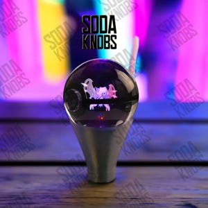May include: A black and silver soda knob with a clear, spherical top. The top features a graphic design and a small red light. The text "SODA KNOBS" is visible in the background.
