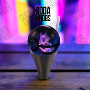 Könnte beinhalten: Ein klarer, kugelförmiger Soda-Knauf mit einem 3D-Bild eines Vaporeon im Inneren. Der Knauf sitzt auf einem silberfarbenen Sockel. Der Text "SODA KNOBS" wird in schwarzen Buchstaben über dem Knauf angezeigt. Der Hintergrund ist eine verschwommene Mischung aus Lila und Gelb.
