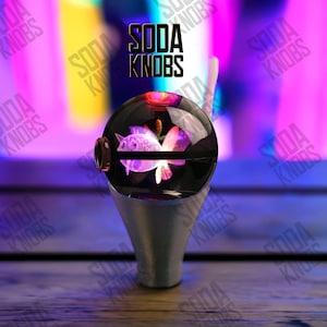 Peut inclure: Un bouton de soda décoratif avec un dessus sphérique transparent présentant un motif de poisson violet. Le bouton repose sur une base argentée. Le texte "SODA KNOBS" est affiché au-dessus du bouton.