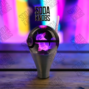 May include: A custom car gear stick knob with a clear, spherical top containing a glowing purple figure. The knob has a silver base and the text "SODA KNOBS" is visible in the background.